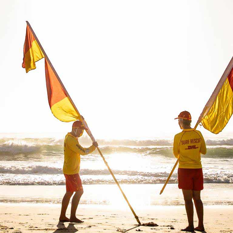 Male surf life saving guard is standing with surf rescue flags on the beach.